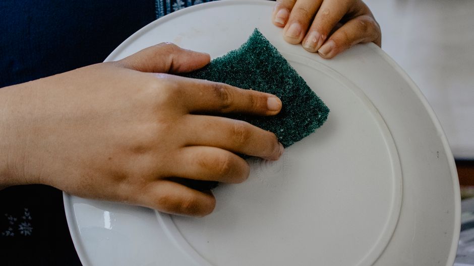 Hands using a green scrub pad to clean a white plate indoors.